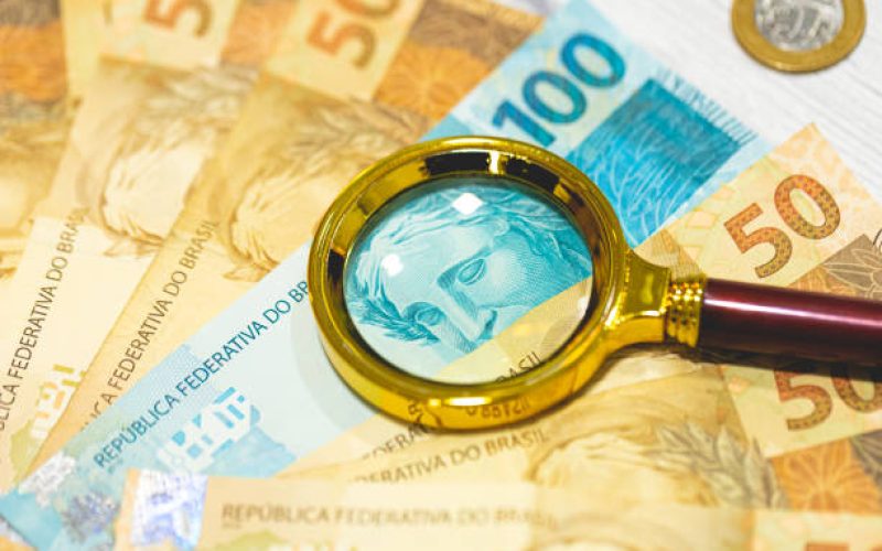 Brazilian Real banknotes on a wooden table with a golden magnifying glass in the composition. Brazilian economy, finance and inflation.