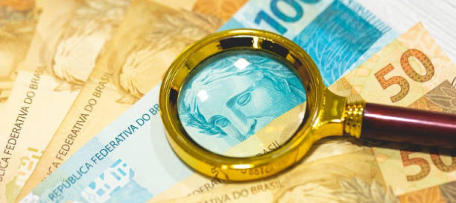 Brazilian Real banknotes on a wooden table with a golden magnifying glass in the composition. Brazilian economy, finance and inflation.